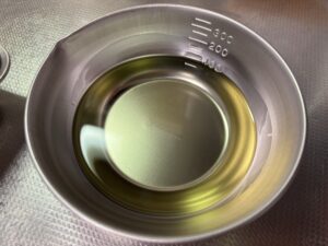 コソリで「揚げ物風」調理をする前に、少量のサラダ油（またはオリーブオイル）を計量カップに用意した様子。 オイルスプレーではなく、食材を油に「くぐらせる」ことで、衣全体にまんべんなく油をコーティングし、ムラなくカリッと仕上げるための工夫です。使用する油の量を最小限に抑えつつ、揚げ物のような満足感を引き出します。